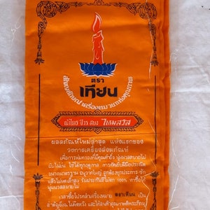 May include: A bright orange fabric package with a black border and text in Thai. The text reads "ตราเครื่องหมา ผ้าไตร จีวร สบง ไหมสวิส" and includes information about the product.
