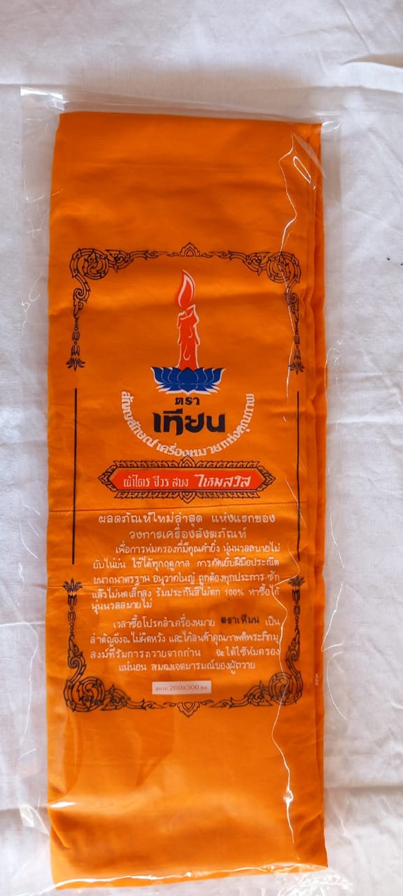 May include: A bright orange fabric package with a black border and text in Thai. The text reads "ตราเครื่องหมา ผ้าไตร จีวร สบง ไหมสวิส" and includes information about the product.