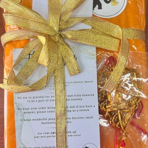May include: An orange package wrapped with a gold ribbon and bow. A gold pendant with a red tassel hangs from the package. The package has a white sticker with a black elephant logo and the text "LANKA MEGAMART".