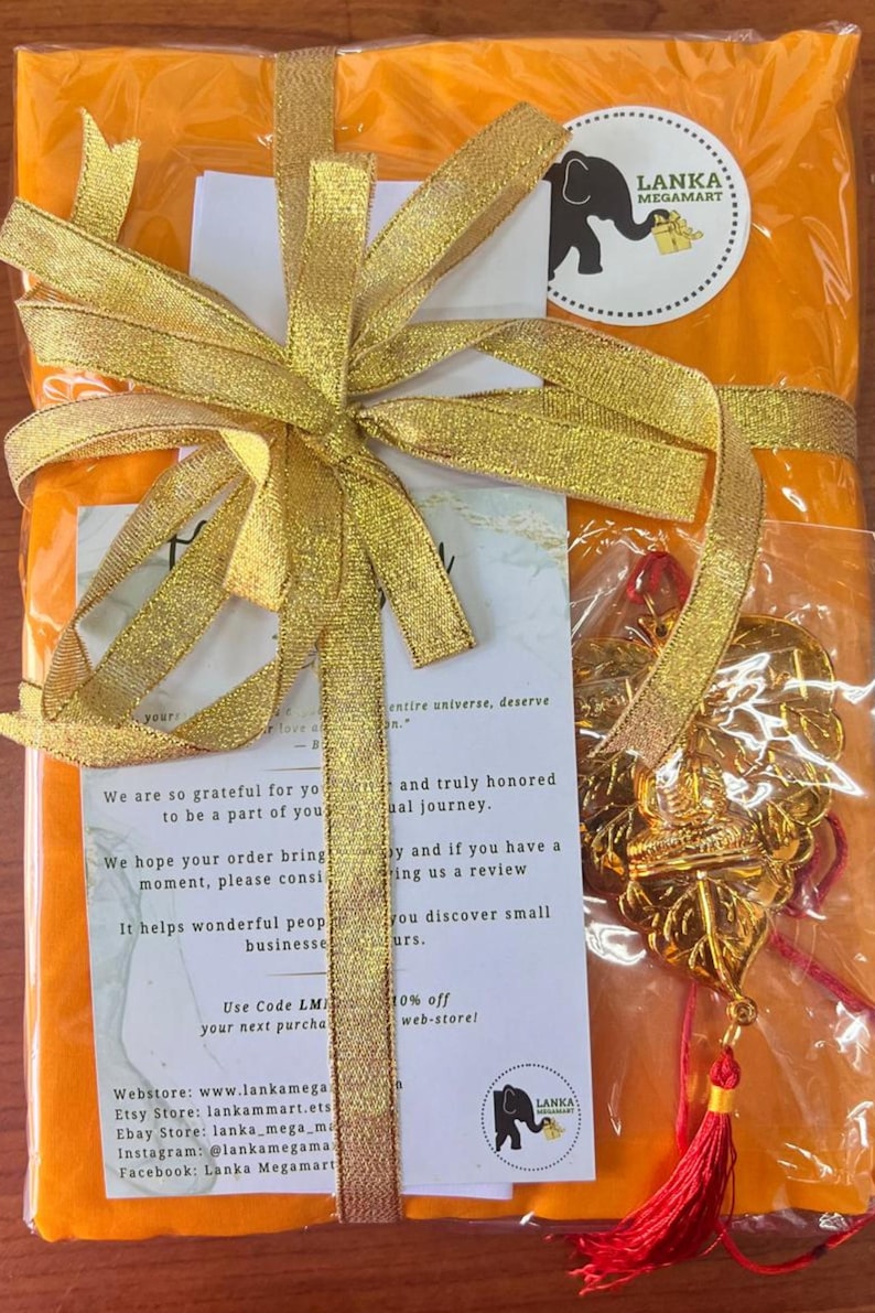 May include: An orange package wrapped with a gold ribbon and bow. A gold pendant with a red tassel hangs from the package. The package has a white sticker with a black elephant logo and the text "LANKA MEGAMART".