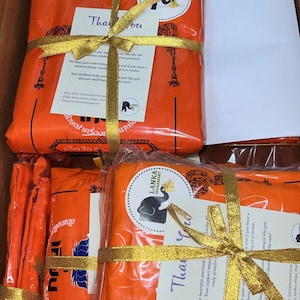 May include: A stack of orange fabric wrapped in gold ribbon and tied with a bow. The fabric is labeled with the text "LANKA MEGAMART" and a small elephant graphic. A thank you card is attached to the ribbon with the text "Thank you" and a message of gratitude.