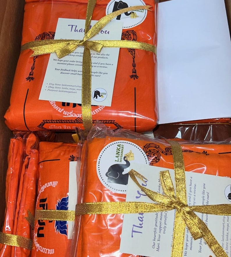 May include: A stack of orange fabric wrapped in gold ribbon and tied with a bow. The fabric is labeled with the text "LANKA MEGAMART" and a small elephant graphic. A thank you card is attached to the ribbon with the text "Thank you" and a message of gratitude.