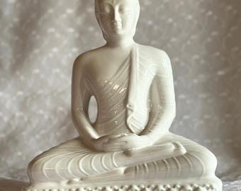 May include: White ceramic statue of Buddha sitting in meditation pose with hands in lap. The statue is on a lotus base.