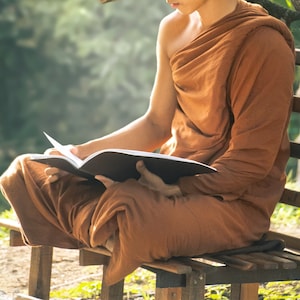 May include: A person wearing a brown robe sits on a wooden bench and reads a book. The book is open and the pages are visible.