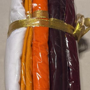 May include: A bundle of folded fabric in white, yellow, orange, and burgundy, wrapped in clear plastic. A gold ribbon with a bow secures the bundle. The fabrics appear to be of a medium weight.