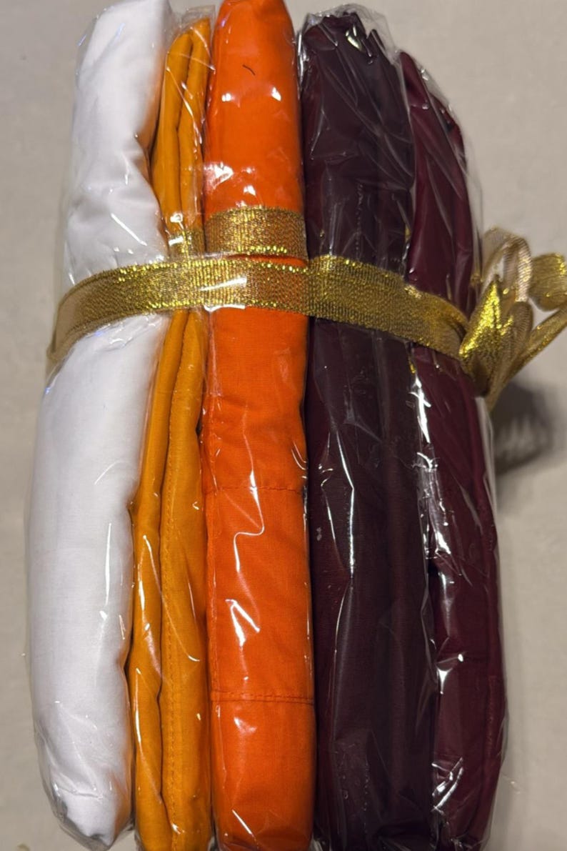 May include: A bundle of folded fabric in white, yellow, orange, and burgundy, wrapped in clear plastic. A gold ribbon with a bow secures the bundle. The fabrics appear to be of a medium weight.