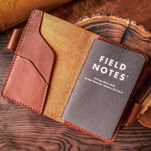 Könnte beinhalten: Ein beigefarbener Notizbuchumschlag aus Leder mit sichtbaren Nähten, einer Seitentasche und einer Stiftschlaufe. Im Inneren befindet sich ein dunkelgraues 48-seitiges Field Notes Notizbuch mit dem Text Field Notes, 48-Page Memo Book.