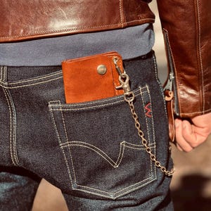 Può includere: Un portafoglio in pelle marrone con una catena d'argento attaccata, parzialmente visibile nella tasca posteriore di jeans blu scuro. I jeans hanno cuciture chiare e un accento rosso. Una giacca di pelle marrone è visibile sullo sfondo.