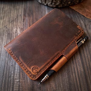 Peut inclure: Une couverture de carnet rectangulaire en cuir marron foncé avec des coutures visibles sur les bords, un logo circulaire gaufré dans le coin inférieur et un passant pour stylo intégré contenant un porte-mine noir à pointe métallique.