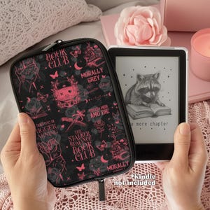 May include: A black Kindle case with pink accents and the words "Book Club" and "Morally Grey" is held next to a Kindle e-reader displaying a raccoon reading a book. The case features a zipper and various graphic designs.
