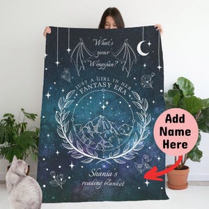 May include: A blue and black reading blanket with a white design featuring a crescent moon, stars, a mountain range, and a wreath. The text "What's your Wingspan? Just a girl in her Fantasy Era" is printed on the blanket. The text "Shania's reading blanket" is printed at the bottom.