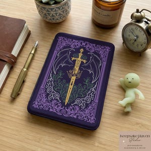 May include: A dark purple journal with a gold sword and crown design, framed by wings and floral elements. The journal is surrounded by a pen, a small clock, a candle, and a small figurine. The text "Keepsake Haven Studio" is visible.