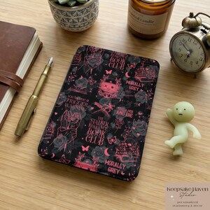 May include: A black notebook with a pink and gray floral and text design, including the words "Stalker Romance Book Club" and "Morally Grey." The notebook is surrounded by a pen, a candle, a small clock, and a small figurine.