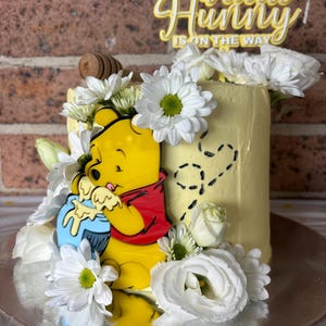Könnte beinhalten: Ein gelber Kuchen, verziert mit einem Winnie-Puuh-Design, einem Honiglöffel und weißen Blumen. Der Kuchen trägt den Text "A little Hunny is on the way" oben. Der Kuchen steht auf einer silbernen Kuchenplatte.