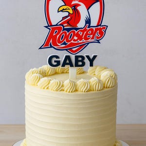 May include: A round, frosted cake with a decorative top. The cake is topped with a Sydney Roosters logo and the name "Gaby". The cake has a yellow frosting with decorative swirls around the top and bottom. The number "40" is also on the cake.