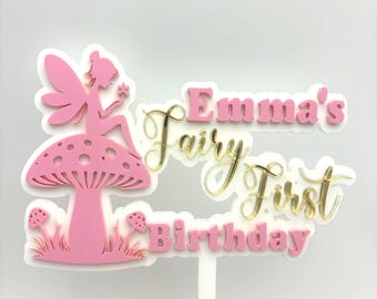 May include: A pink and white cake topper featuring a fairy sitting on a mushroom with the text Emma's Fairy First Birthday in pink and gold script and block lettering. The design includes small mushrooms at the base and a white support stick.