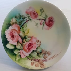 Puede incluir: Un plato redondo de color crema con un diseño pintado a mano de rosas rosas con hojas verdes y tallos espinosos. Las rosas están agrupadas a la izquierda, sobre un fondo suave y difuminado de color verde y amarillo.