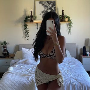 Peut inclure: Une femme porte un haut de bikini à imprimé léopard et une jupe en crochet crème. La jupe est nouée sur le côté. L'arrière-plan comprend un lit avec une literie blanche, une image encadrée et des objets décoratifs sur une étagère.