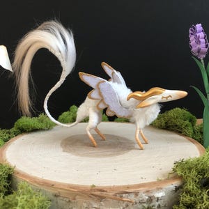 May include: A small, four-legged creature figurine with white fur, gold-colored horns, and white wings with gold trim. It features a long, thin tail with a tuft of white hair at the end and closed eyes. The creature stands in a profile position.