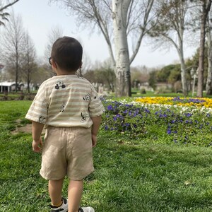 May include: A young child wearing a striped beige and brown shirt with a cartoon character print and beige shorts stands in a grassy park with flowers in the background.