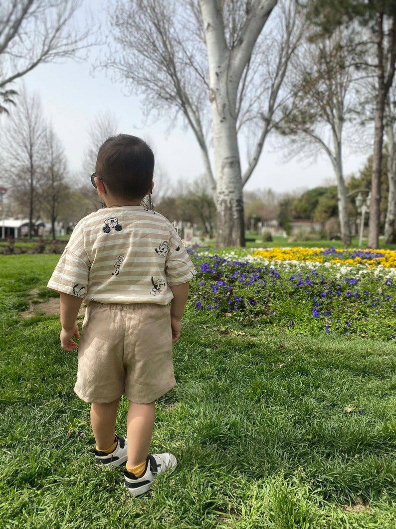 May include: A young child wearing a striped beige and brown shirt with a cartoon character print and beige shorts stands in a grassy park with flowers in the background.