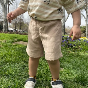May include: A young child wearing a striped beige t-shirt with a cartoon character print and beige shorts. The child is wearing white trainers with black accents and socks with a brown and yellow pattern. The child is standing on a grassy surface.