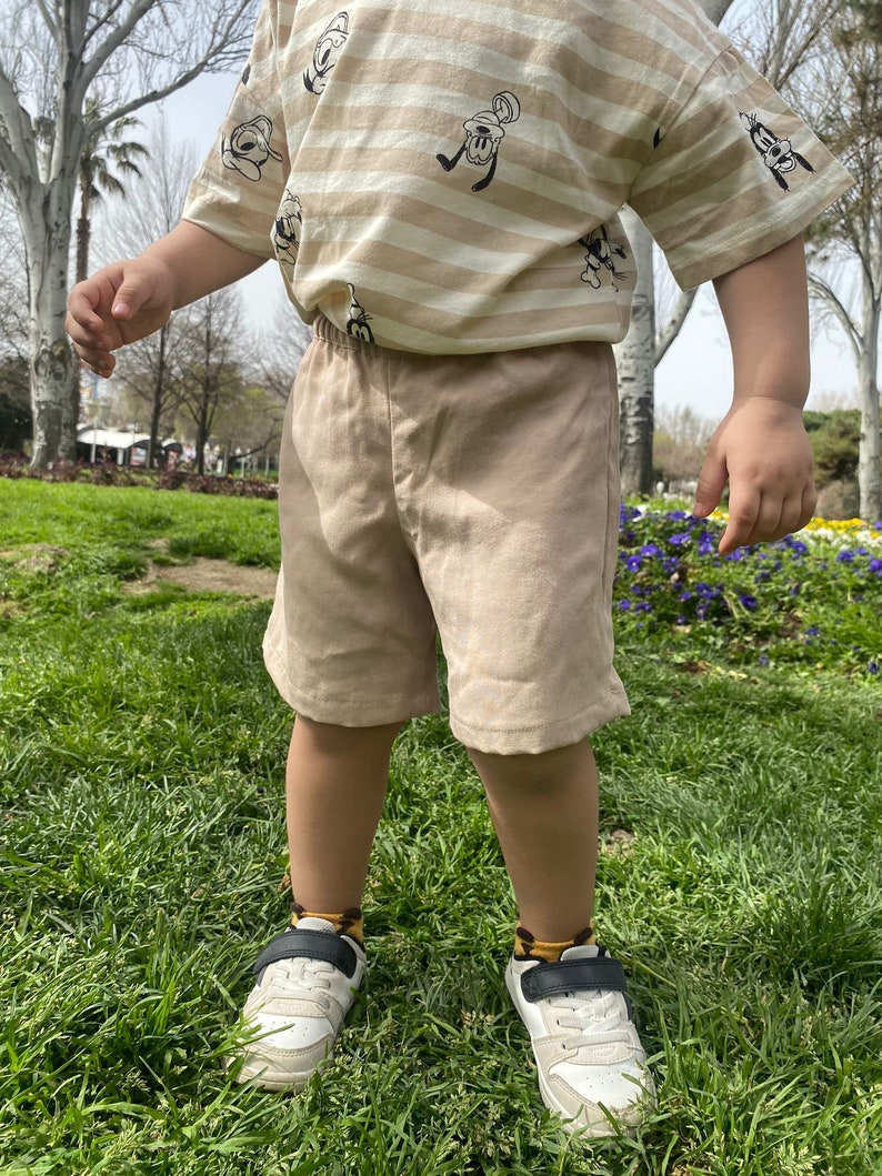 May include: A young child wearing a striped beige t-shirt with a cartoon character print and beige shorts. The child is wearing white trainers with black accents and socks with a brown and yellow pattern. The child is standing on a grassy surface.