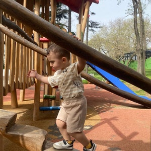 May include: A young child wearing a striped shirt and khaki shorts climbs a wooden playground structure. The child is holding onto a wooden beam with one hand and has their other hand raised in the air. The playground is made of natural wood and has a blue slide in the background.