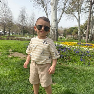 May include: A young boy wearing a striped shirt with cartoon characters, khaki shorts, and sunglasses. He is standing in a grassy area with flowers in the background.