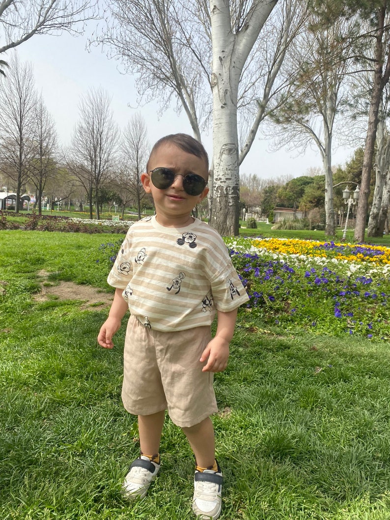May include: A young boy wearing a striped shirt with cartoon characters, khaki shorts, and sunglasses. He is standing in a grassy area with flowers in the background.
