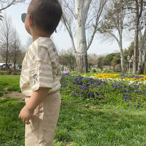 May include: A young child wearing a striped tan and white t-shirt with a cartoon graphic and tan shorts stands in a park. The child is wearing sunglasses and looking up at the sky.