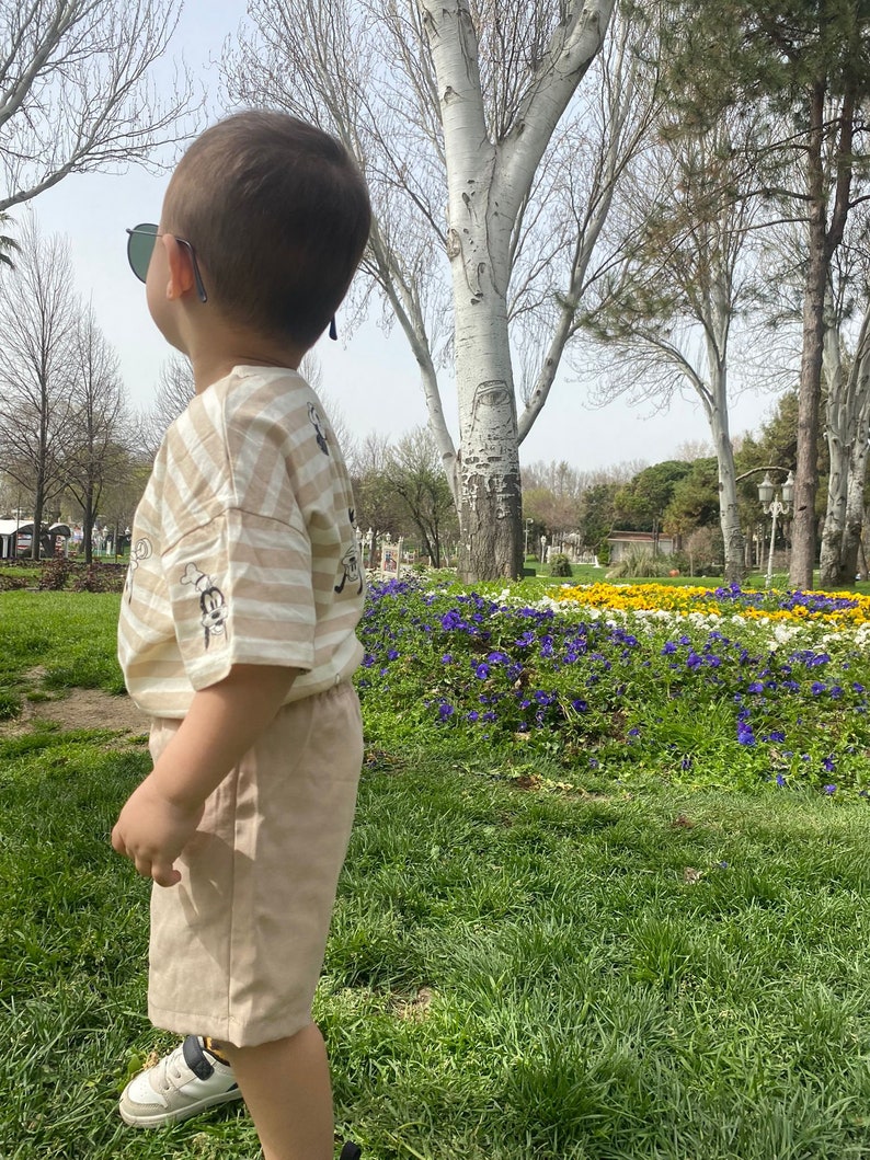 May include: A young child wearing a striped tan and white t-shirt with a cartoon graphic and tan shorts stands in a park. The child is wearing sunglasses and looking up at the sky.
