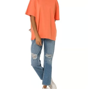 May include: A coral colored oversized t-shirt with short sleeves and a relaxed fit. The model is wearing the t-shirt with blue distressed jeans and white sneakers.