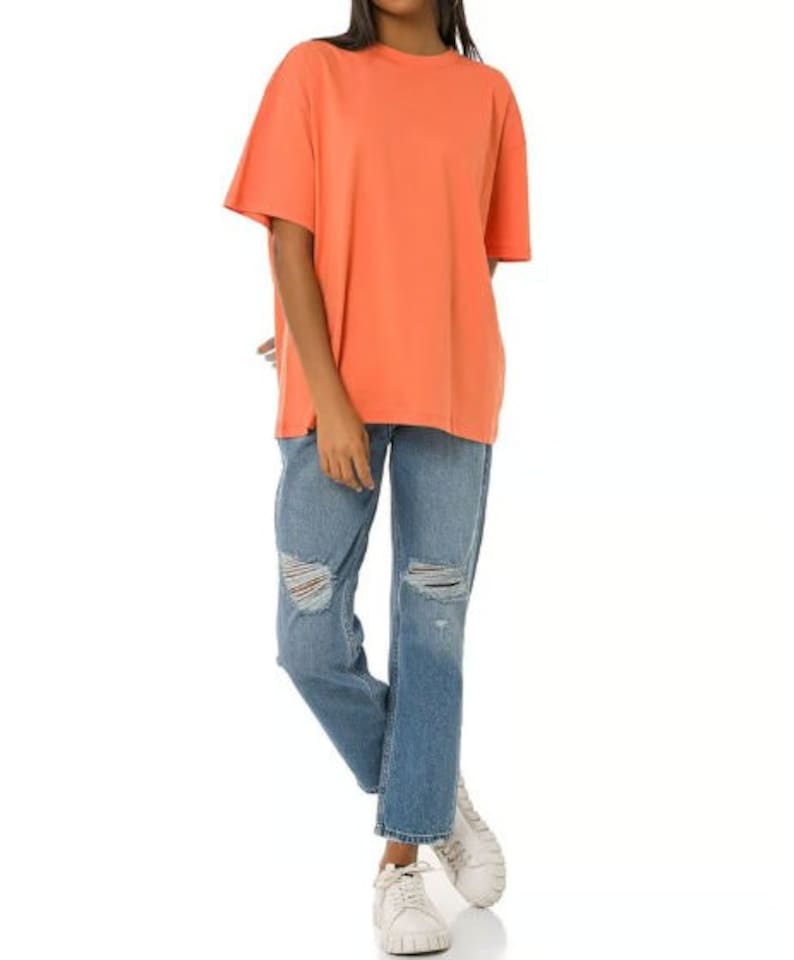 May include: A coral colored oversized t-shirt with short sleeves and a relaxed fit. The model is wearing the t-shirt with blue distressed jeans and white sneakers.