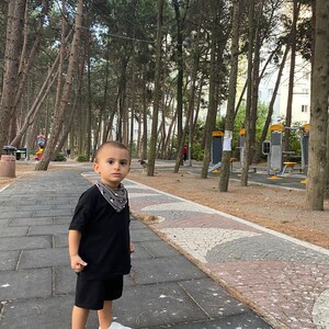 May include: A young boy wearing a black t-shirt and black shorts stands on a paved path in a park. He is looking at the camera. The path is surrounded by trees.
