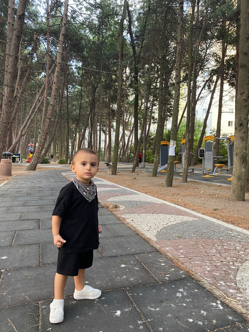 May include: A young boy wearing a black t-shirt and black shorts stands on a paved path in a park. He is looking at the camera. The path is surrounded by trees.