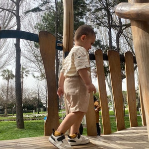 May include: A young child wearing a beige and white striped shirt and beige shorts stands on a wooden platform. The child is wearing white trainers with black accents and is looking down. The platform is surrounded by wooden railings.
