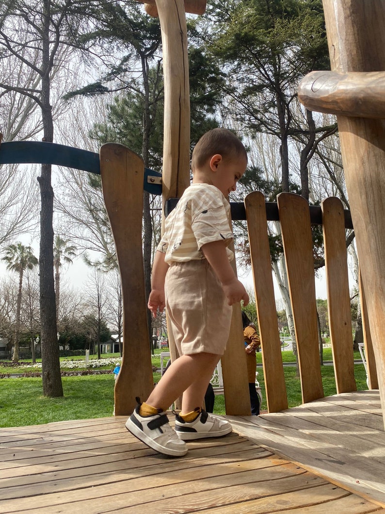 May include: A young child wearing a beige and white striped shirt and beige shorts stands on a wooden platform. The child is wearing white trainers with black accents and is looking down. The platform is surrounded by wooden railings.