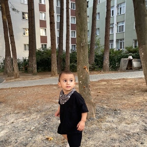 May include: A young boy wearing a black t-shirt and black shorts stands in a park. He is wearing a bandana with a black and white pattern around his neck. He is looking to the right of the frame.