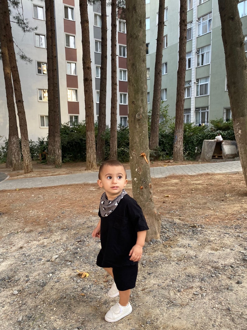 May include: A young boy wearing a black t-shirt and black shorts stands in a park. He is wearing a bandana with a black and white pattern around his neck. He is looking to the right of the frame.