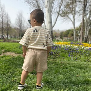 May include: A young child wearing a striped shirt with a cartoon character print, beige shorts, and black trainers stands in a grassy park with flowers in the background.