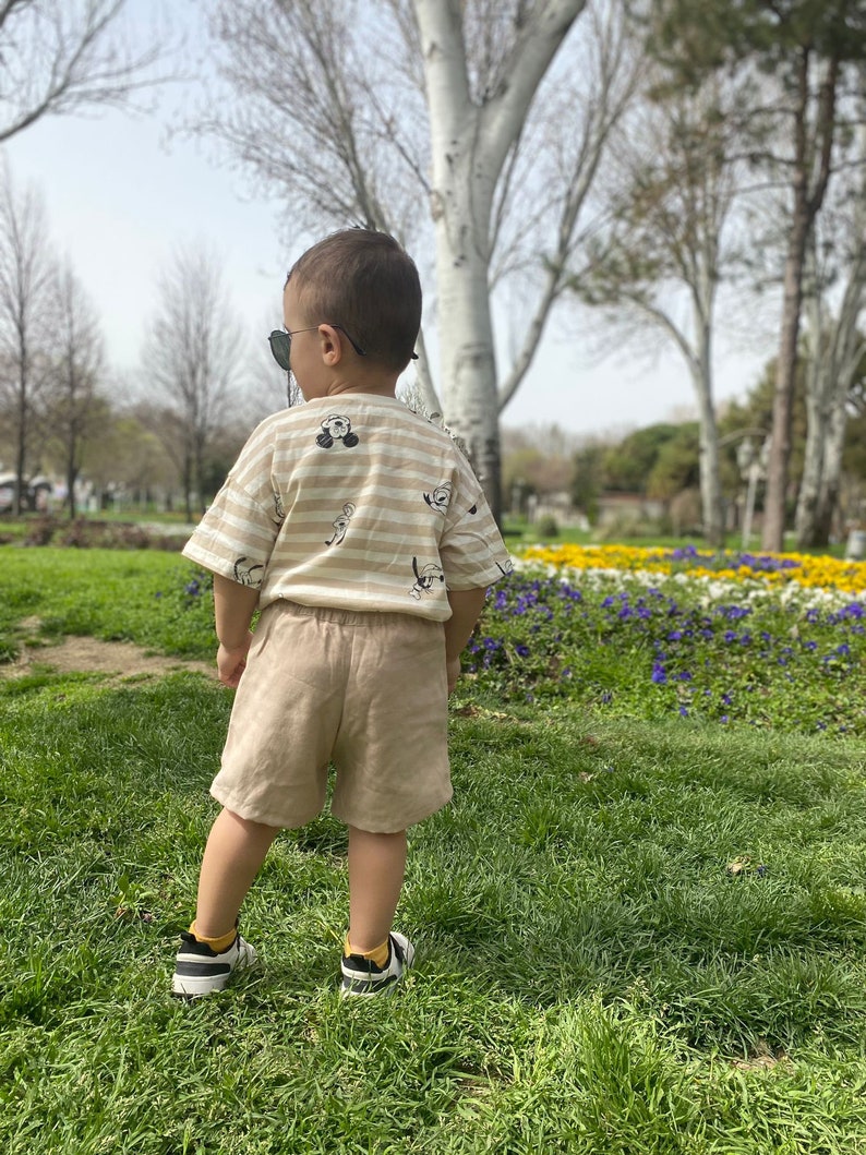 May include: A young child wearing a striped shirt with a cartoon character print, beige shorts, and black trainers stands in a grassy park with flowers in the background.