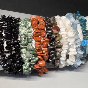 May include: A collection of seven stretch bracelets made from irregular, tumbled gemstone chips. The bracelets feature distinct colors including black, mottled green, shimmering orange-brown, dark gray, white, pale blue, and deep teal blue.
