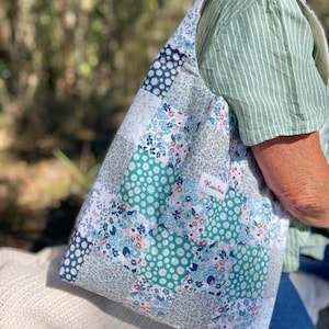 May include: A hobo-style shoulder bag featuring a patchwork design of various floral and geometric patterns in shades of teal, navy blue, white, and soft pink. A small white rectangular fabric label with the text Rowbie is sewn onto the front.