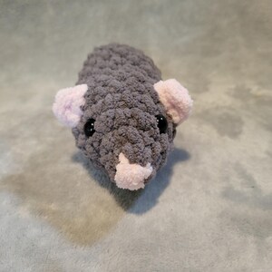 May include: A handmade gray crochet rat with pink ears and nose. The amigurumi rat has black safety eyes and is made with a soft, textured yarn. The plush toy is a cute and cuddly creation.