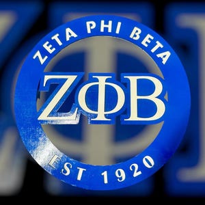 Könnte beinhalten: Ein blau-weißes Zeta Phi Beta-Schild. Das kreisförmige Schild zeigt die griechischen Buchstaben ZΦB in der Mitte, umgeben von den Worten "ZETA PHI BETA" und "EST 1920". Der Text ist weiß auf blauem Hintergrund.
