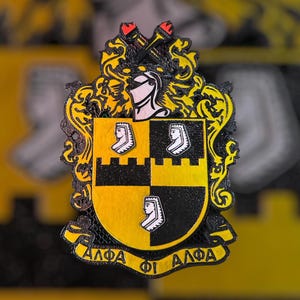 Peut inclure: Un emblème décoratif aux couleurs noir et jaune. L'emblème présente un motif de bouclier avec trois portraits de profil blancs sur un fond jaune et noir. Le haut de l'emblème comporte un casque de chevalier et deux torches. Les lettres grecques "ΑΛΦΑ ΦΙ ΑΛΦΑ" sont en bas.