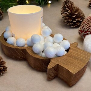May include: A wooden candle holder shaped like a snowman, filled with white spheres and a frosted glass candle. Pinecones and a white scarf with red accents are in the background. A festive, winter-themed decoration.