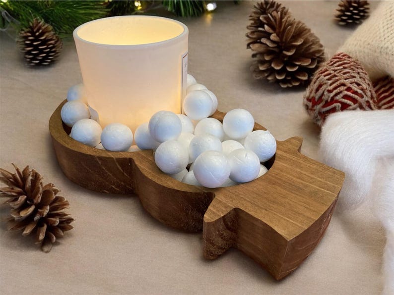May include: A wooden candle holder shaped like a snowman, filled with white spheres and a frosted glass candle. Pinecones and a white scarf with red accents are in the background. A festive, winter-themed decoration.