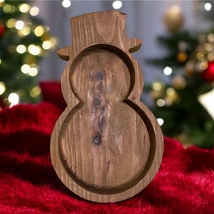 May include: A wooden snowman-shaped tray with a hat. The tray is made of natural wood and has a carved design. The tray is sitting on a red surface, with a Christmas tree in the background.